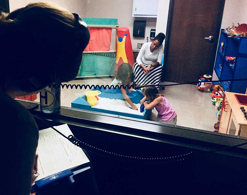 A participant in Play Therapy training peers through a two-way mirror as a counselor observes a child playing as part of therapy.